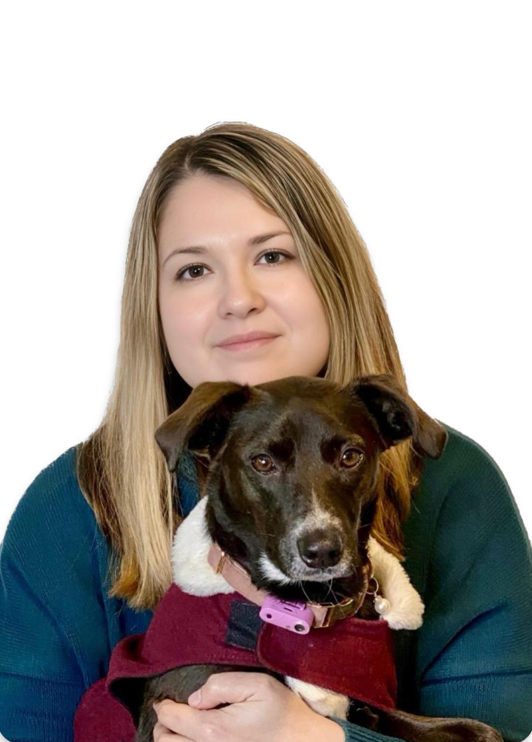 Elena Grafton holding a black and white dog dressed in a maroon outfit with a white collar. Both are looking directly at the camera. Elena has long, straight blonde hair and is wearing a teal sweater