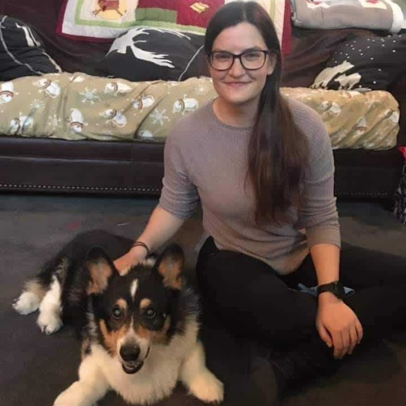 Laura Muzal, a woman with long dark hair and glasses, wearing a grey sweater, sits on the floor with a black, white, and brown Corgi dog. She is smiling at the camera, and the dog is lying down, also looking at the camera. Behind them is a couch with festive blankets and pillows.
