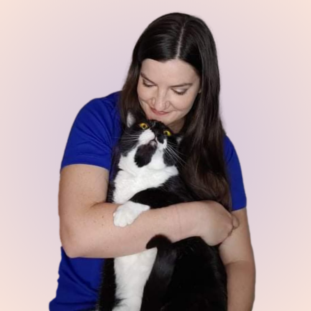 Laura Muzal, a woman with long dark hair, wearing a blue top, is holding a black and white cat. She looks down lovingly at the cat, which has bright yellow eyes. The background is a soft gradient of light colors.