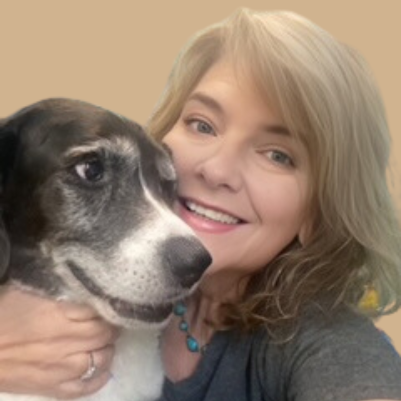 Lori Gammon, a woman with shoulder-length blonde hair, is smiling and hugging a black and white dog. She is wearing a gray top and a turquoise necklace. The background is a plain tan color.