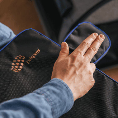 Close-up image of a person's hand holding a PetsPEMF pad, which is black with blue trim. The person's hand, wearing a blue shirt, is visible, and they are lifting a flap on the pad. The PetsPEMF logo is printed on the pad in white. The background is slightly blurred, showing parts of furniture and the floor.