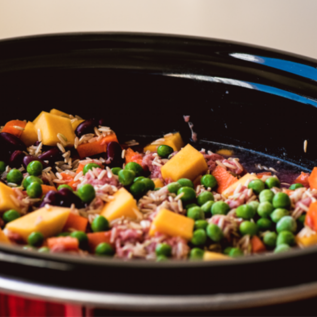 The image shows a black crockpot with a colorful mix of ingredients: diced yellow squash or pumpkin cubes, bright green peas, dark red kidney beans, brown rice with shredded meat (likely ham or corned beef), and chopped carrots. The dish looks vibrant and appetizing, and the clean, shiny crockpot indicates the meal is still in the early stages of cooking.