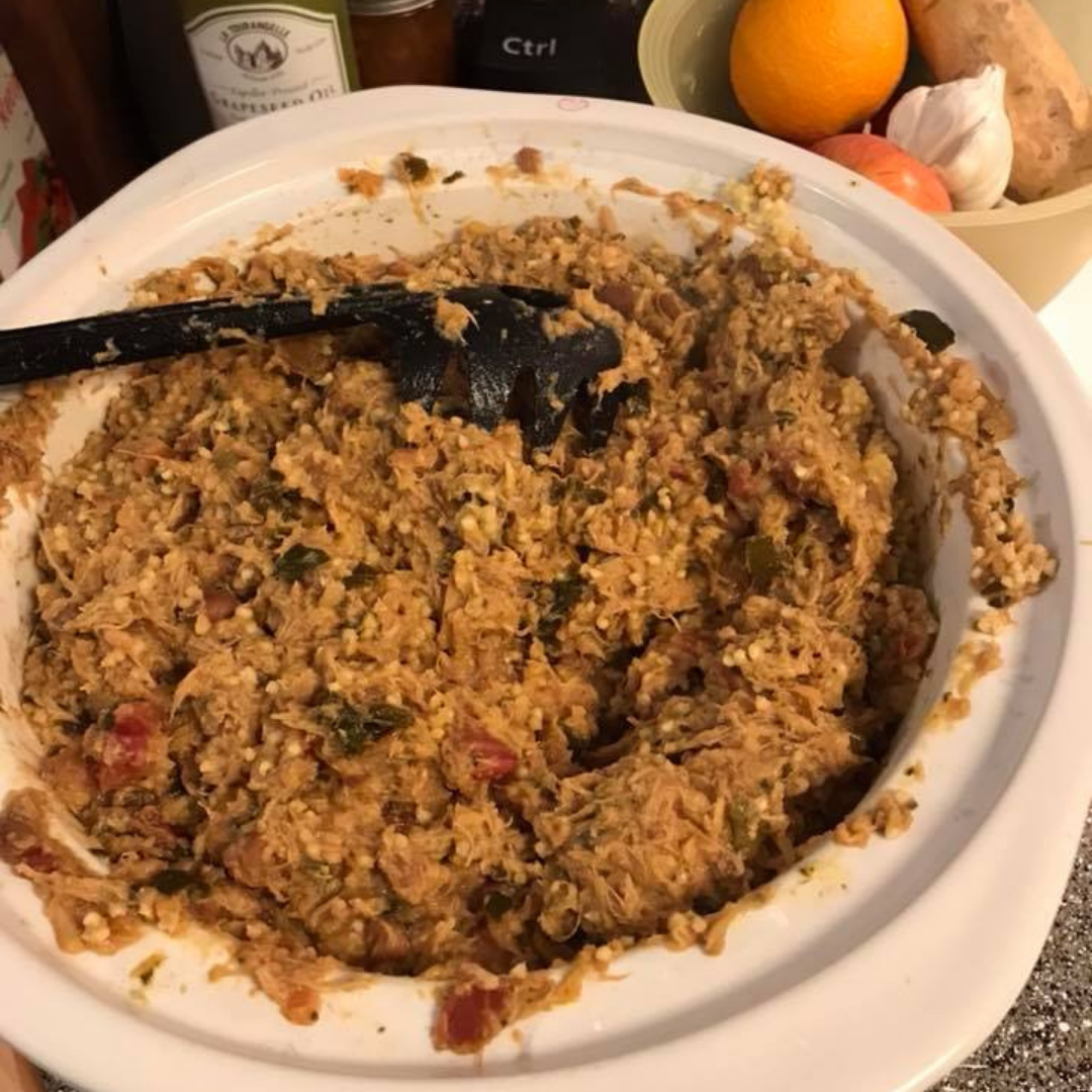 A close-up image of a large bowl filled with a mixture of home-cooked pet food. The mixture includes ingredients like shredded meat, vegetables, and grains. A black serving spoon is placed in the bowl. In the background, there are various kitchen items, including a bottle of grapeseed oil and a bowl of fruits and vegetables.