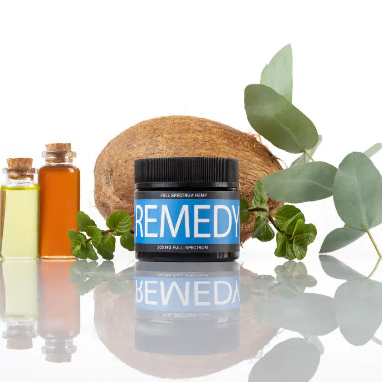 A jar of Full Spectrum Hemp REMEDY for dogs placed in front of a coconut and surrounded by green leaves and small bottles of oils. The jar has a blue label with white text reading 'FULL SPECTRUM HEMP' and 'REMEDY.' It also indicates '300 MG FULL SPECTRUM' at the bottom.